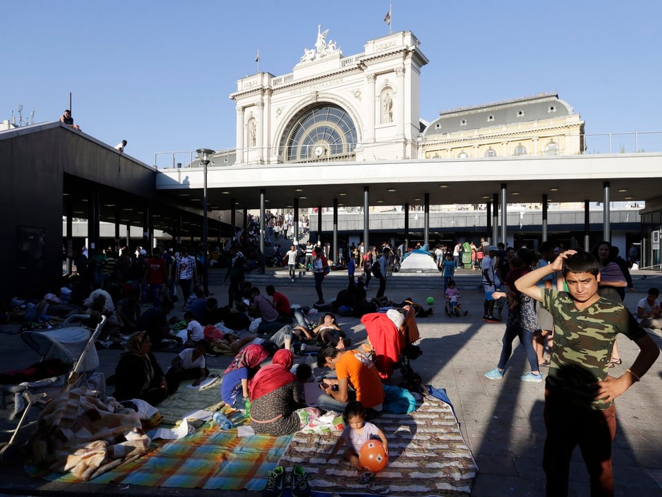 Ein Platz vor dem Bahnhofsgebäude: Familien sitzen auf Decken am Boden, ein Junge (rechts) steht.