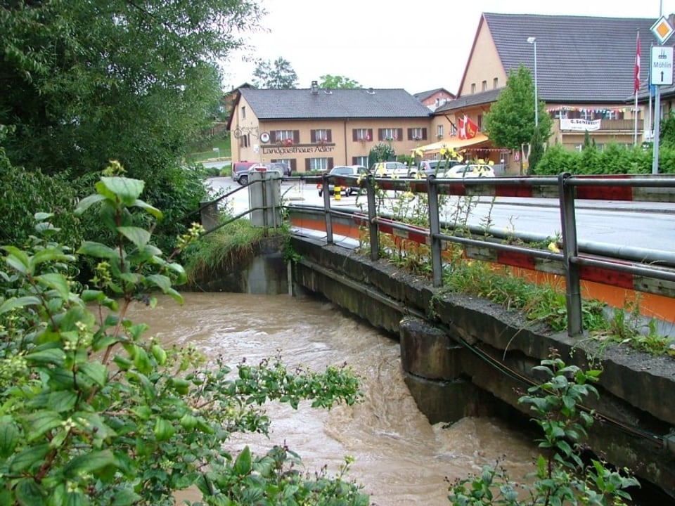 Bach ist braun gefärbt und füllt fast das ganze Bachbett aus, so dass das Wasser nur noch knapp unter der Strassenbrücke hindurch kann.