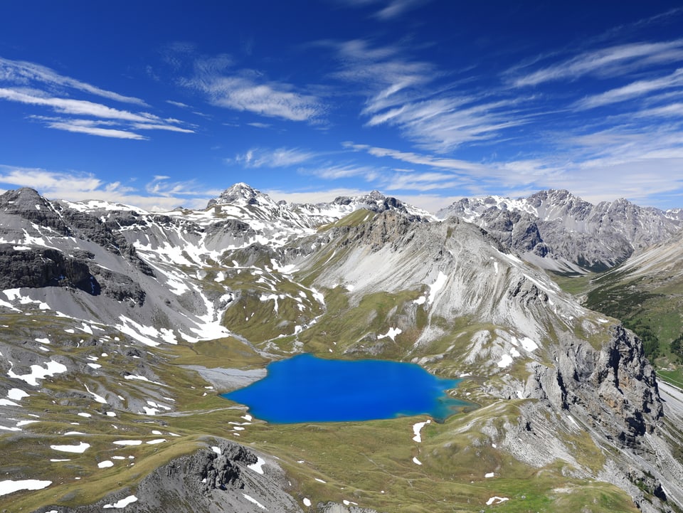 Von einem Gipfel blickt man auf eine Alp hinunter. Ein kleiner Bergsee leuchtet im selben Blau wie der Himmel. Ein paar Schleierwolken schmücken den Himmel.