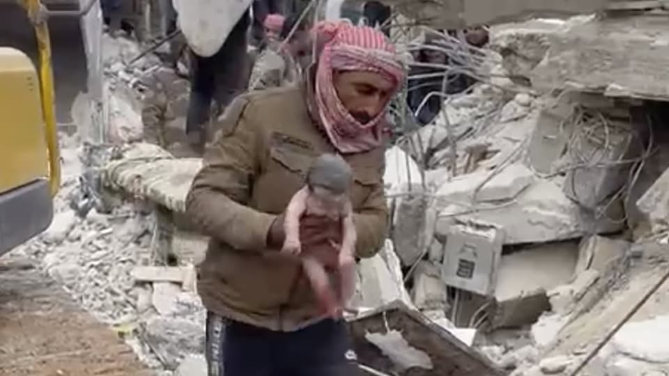 Ein Mann mit Kopftuch trägt ein Baby an Trümmern vorbei
