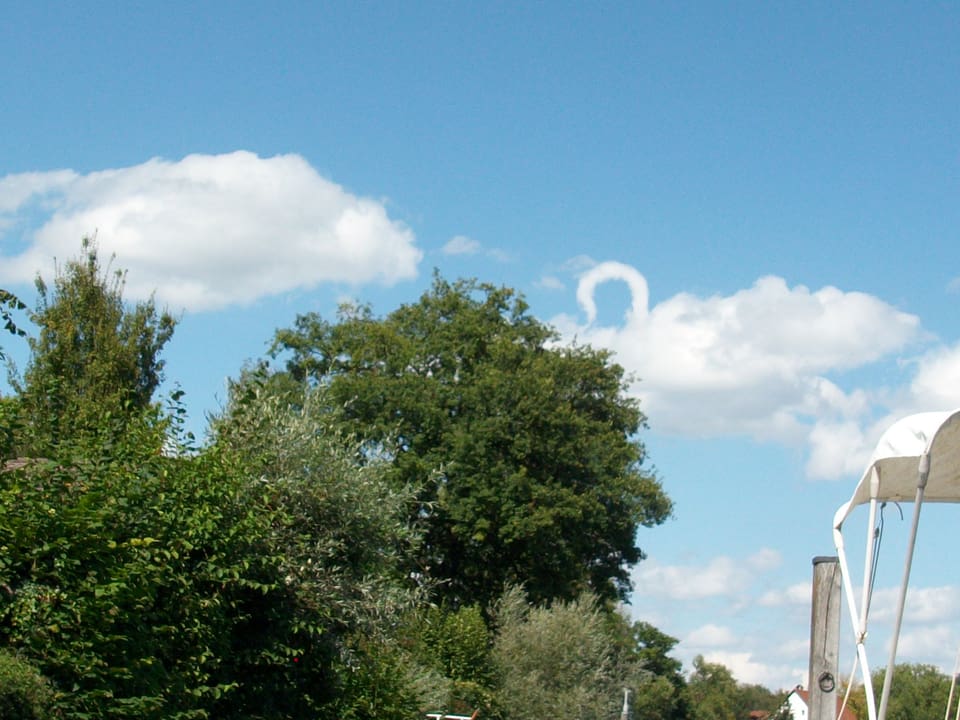 Am blauen Himmel zeigen sich ein paar Wolken. Eine Wolken besitzt am linken Ecken oberhalb der Wolken einen weissen Bogen. Unten links im Bild sind zudem Bäume und Büsche und rechts ein Bootsdach auszumachen.