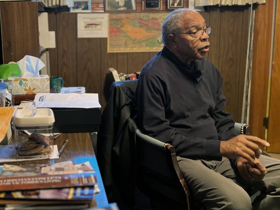 älterer schwarzer Mann mit Brille sitzt in Büro mit Unterlagenstapel, schaut nach links.