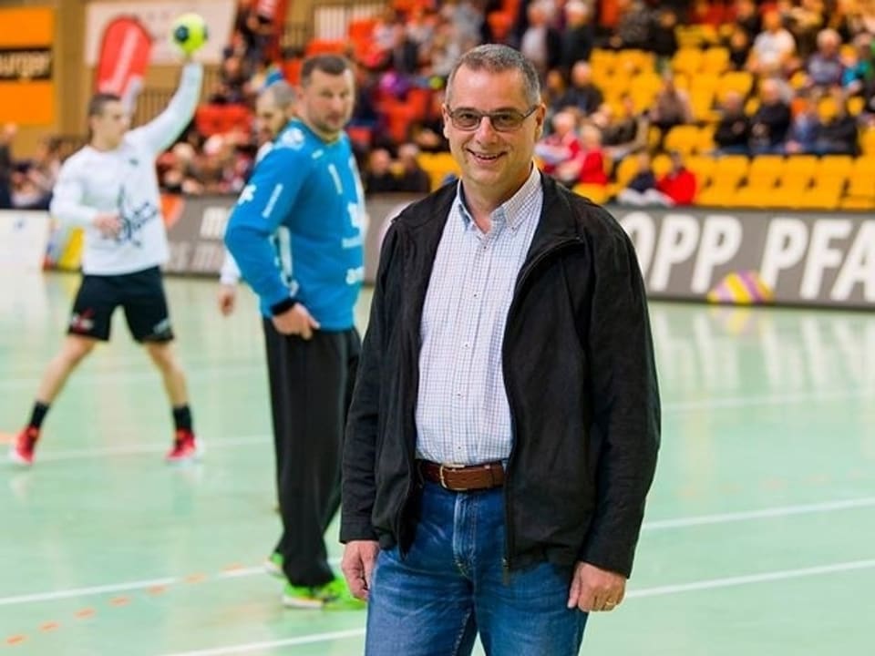 Ein Mann mit Brille steht in einer Turnhalle. Im Hintergrund wirft ein Handballer einen Ball Richtung Tor.