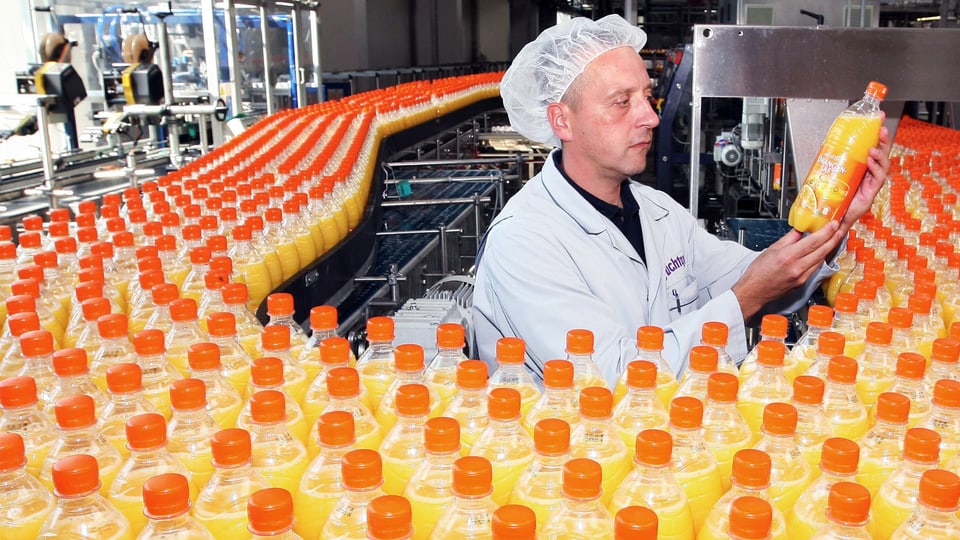 Mann mit Haarnetz steht inmitten einer Orangensaft-Produktionsstrasse und hält eine Flasche prüfend in die Höhe.