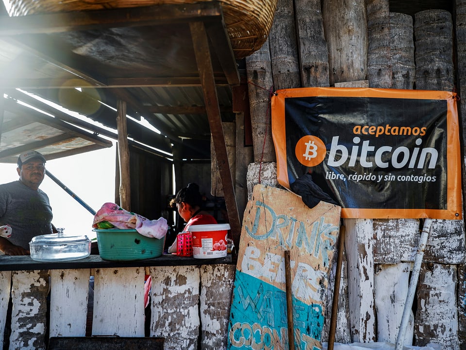 An einem Stand hängt ein Banner, worauf in spanisch steht, dass Bitcoin akzeptiert wird. Man sieht nebendran den Tresen.