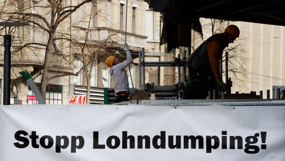 Ein Plakat mit der Aufschrift «Stopp Lohndumping», dahinter zwei Bauarbeiter bei der Arbeit.