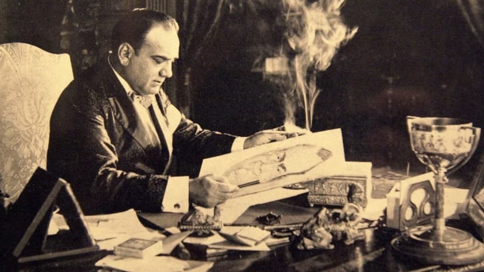 Ein Herr im Anzug sitzt an einem Schreibtisch umgeben von Zigarrenrauch.