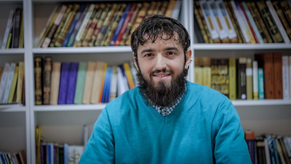 Ardian Elezi schaut freundlich in die Kamera. Er sitzt vor einer Bücherwand, hat einen Bart und trägt ein blauer Pulli.