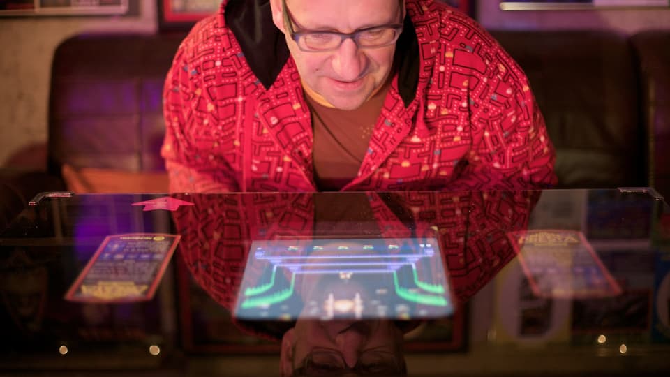 Ein Mann mit roter Pac Man-Jacke sitzt an einem Videospiel-Tisch
