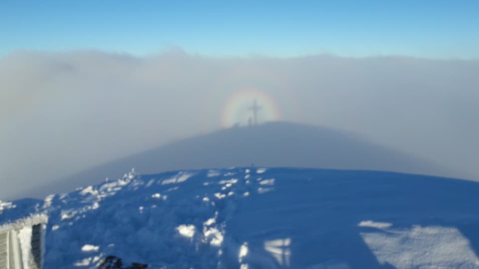 Im Nebelschatten ist ein Mann neben einem Gipfelkreuz zu erkennen. Um den Kopf des Schattens hat sin eine Glorie gebildet.