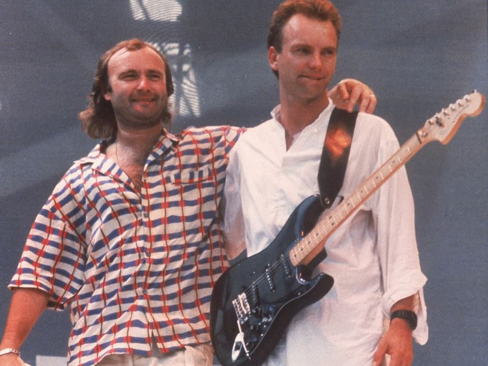 Zwei Männer stehen auf einer Bühne. Einer trägt ein bunter Shirt und umarmt den anderen, der eine Gitarre um den Hals trägt.