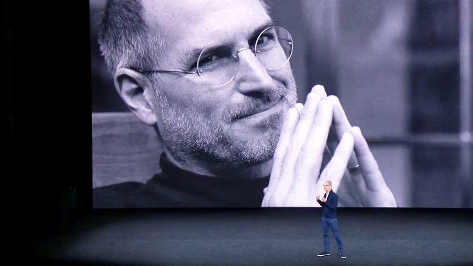 Eine Nahaufnahme von Steve Jobs wird auf eine grosse Wand projiziert. Davor steht ein Mann in Jeans und hält eine Präsentation.