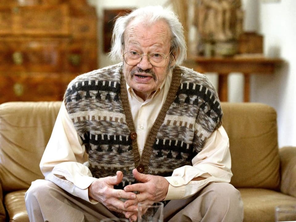 Alter bebrillter Herr in Hemd und braun-grauer Strickjacke sitzt auf Sofa