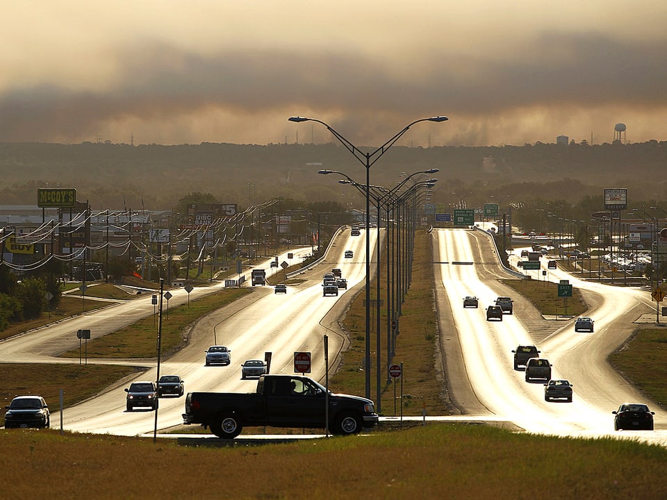 Blick über einen Highway. Die Strassen scheinen zu glühen. Über dem Horizont türmen sich Rauchwolken.
