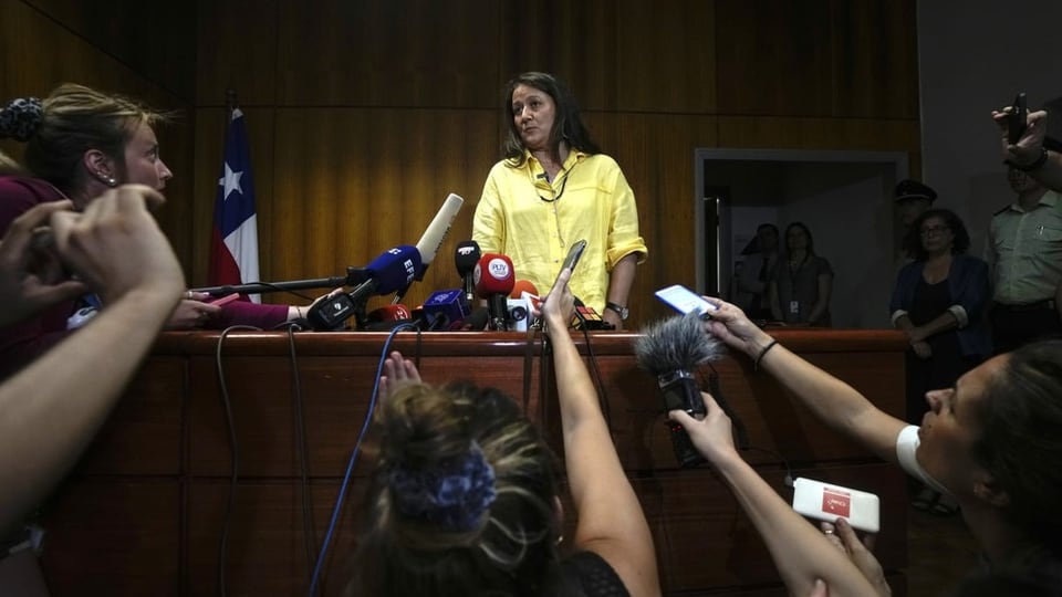 Ein Mittsechzigerin in gelbem Hemd steht vor einer Traube von Journalisten, die ihr Mikros entgegenrecken.