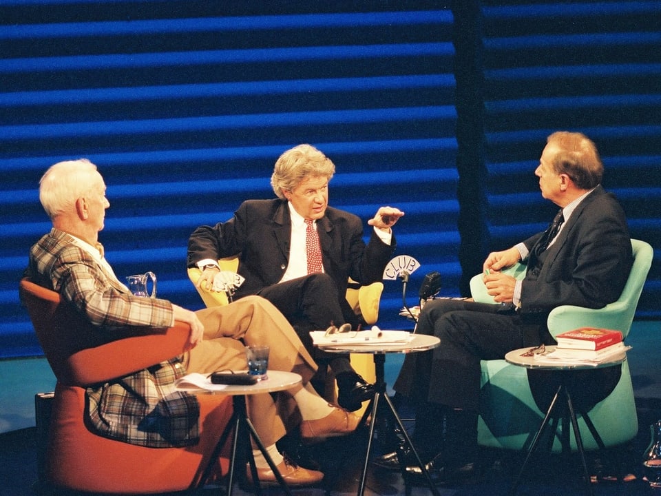 Drei Männer sitzen auf bunten Sesseln und diskutieren.