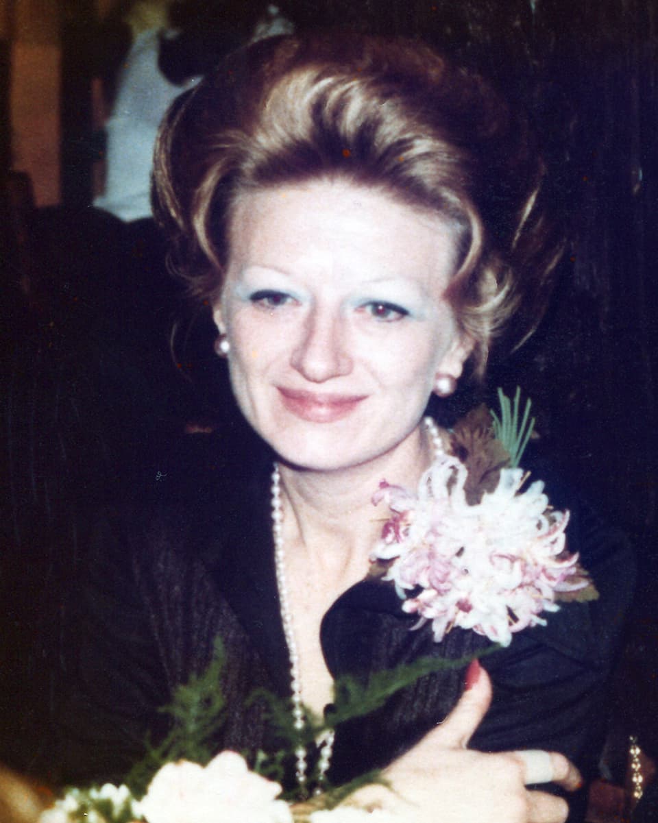 Erna Schürch mit eleganter Frisur und einen grossen Blume am Blusenkragen sitzt an einem Tisch.