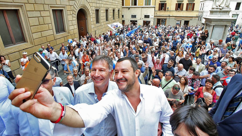 Salvini macht ein Selfie, neben ihm steht ein Mann, beide grinsen in die Kamera, dahinter viele Leute.