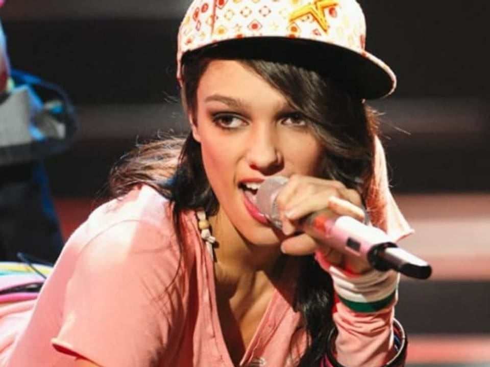 A young woman lies on the floor and sings.  She has a hat and a microphone in her hand.