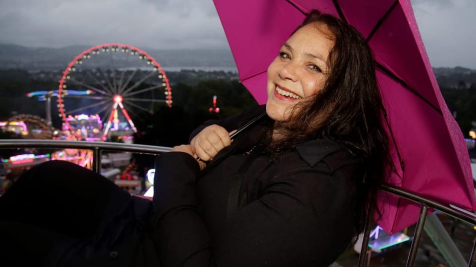 Frau mit schwarzen Haaren sitz lachend mit Schirm in einem Riesenrad