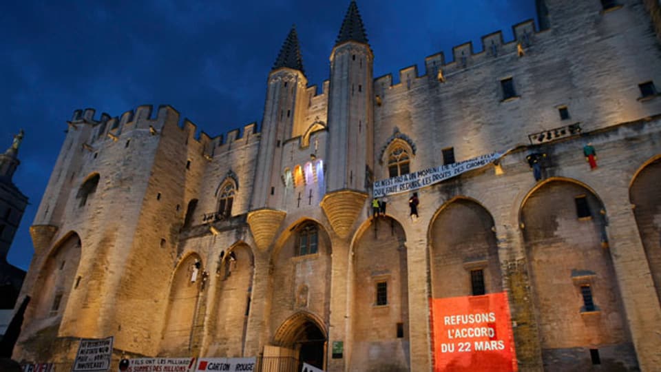 Die Fassade des Papstpalastes in Avignon, beleuchtet bei Nacht. Im Vordergrund Demonstranten und ein Banner mit der Aufschrift: «Refusons l'accord du 22 mars"