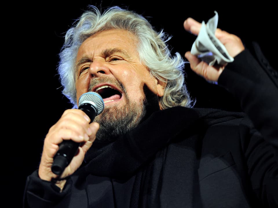 Beppe Grillo während einer Rede mit Mikrofon und einem Zettel in der Hand