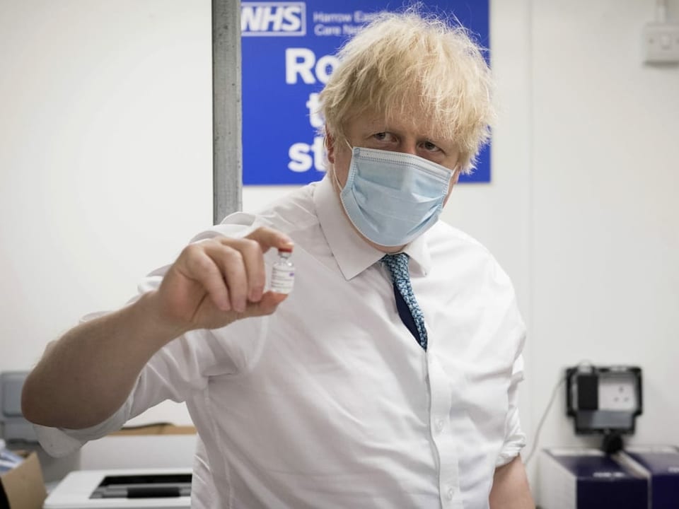 Johnson trägt eine Maske und hält ein Gläschen mit Impfstoff in die Höhe.
