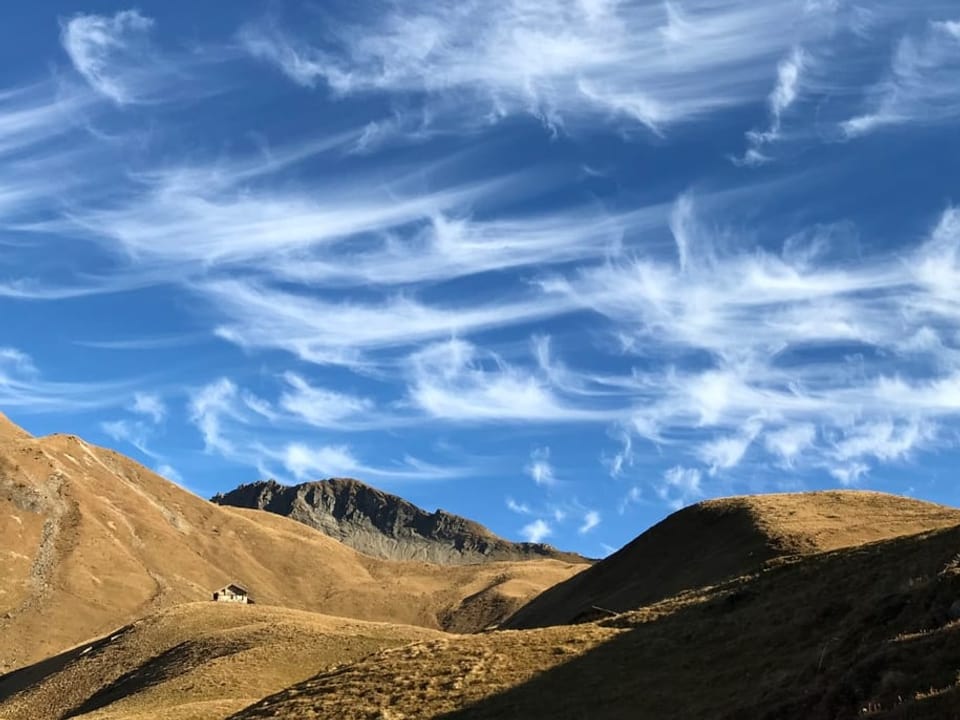 Federwolken, die gebogen sind am tief blauen Himmel über olivfarbenen Berghängen.