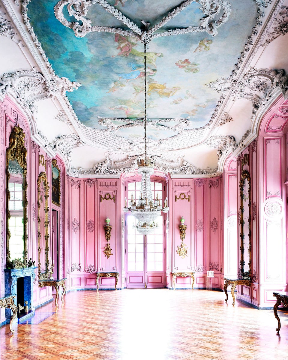 Blick in einen Saal mit Parkettboden, rosafarbenen Wänden mit goldenen Leuchtern und einer reich verzierten weiss-türkisfarbenen Decke, in der Mitte ein Kronleuchter.
