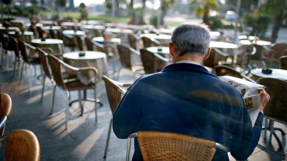 Ein Mann (von hinten) sitzt allein an einem Tisch draussen vor einem Café und liest Zeitung.