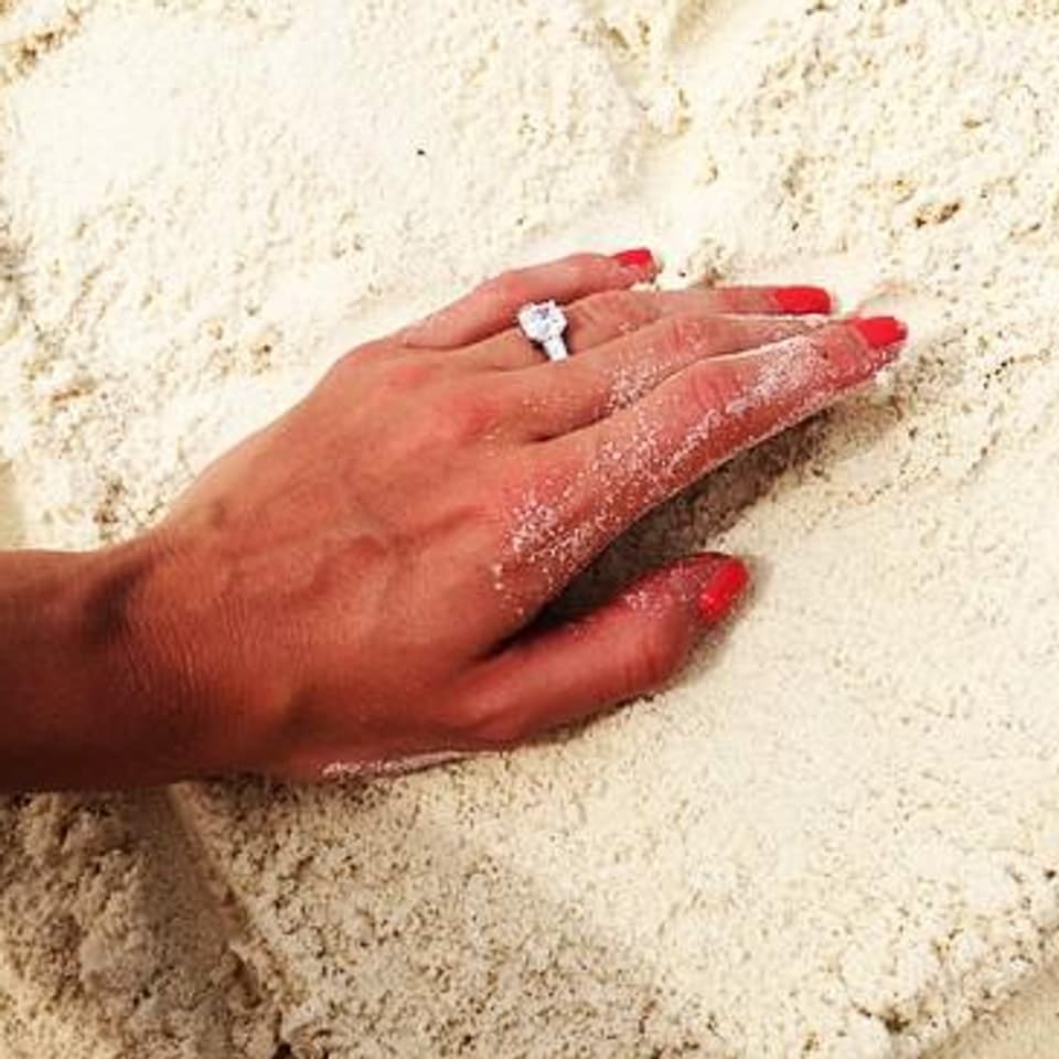 Eine Hand mit Verlobungsring in einem Herz aus Sand. 