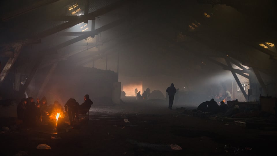 Eine verlassene Industriehalle im Rauch. An mehereren Stellen brennen Feuer, rundum sitzen jeweils ganze Menschengruppen und wärmen sich.