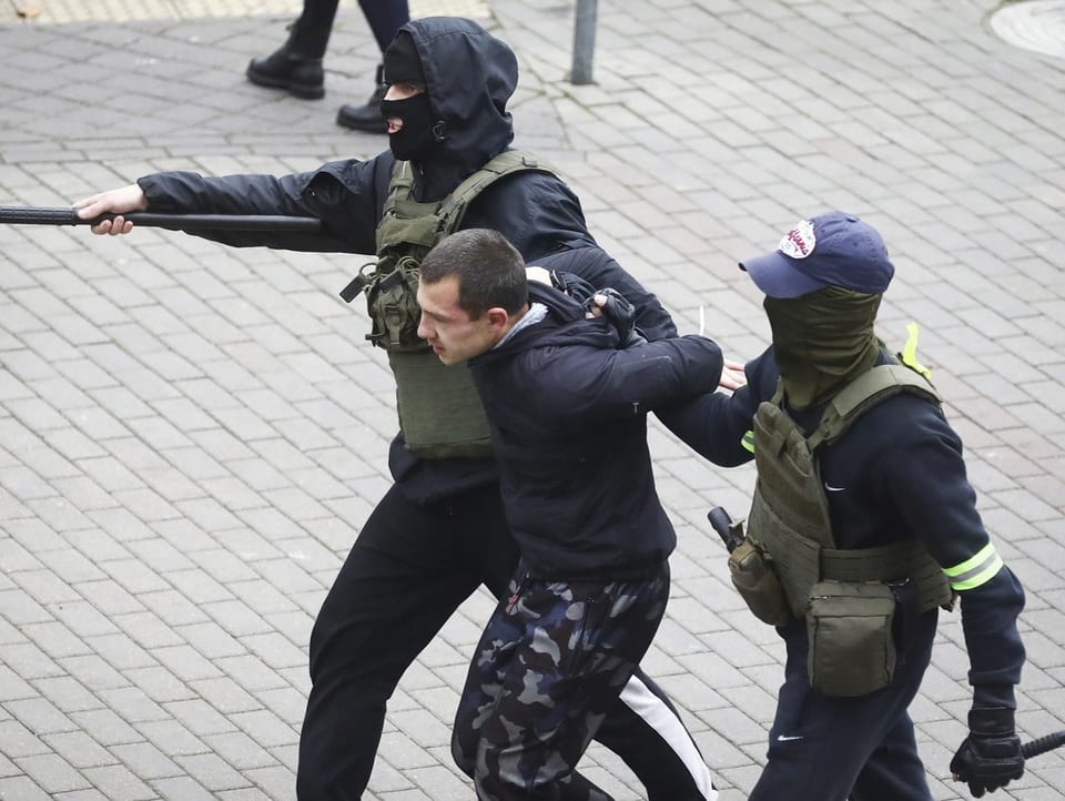 Two masked police officers lead a protester away.  They both carry clubs.