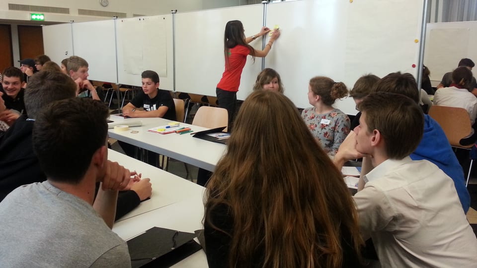 Schüler sitzen an einem Tisch, Frau klebt Zettel an Wandtafel.