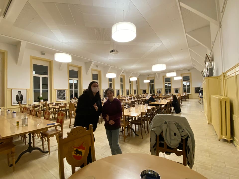 Die beiden Mädchen stehen links im Bild. Im Hintergrund sieht man eine grosse Halle mit leeren Tischen und Stühlen.