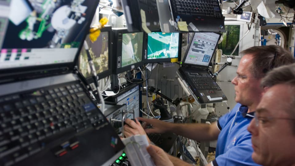 Astronauten an Bord der ISS sitzen vor einer Reihe von an die Wand montierten Laptops.