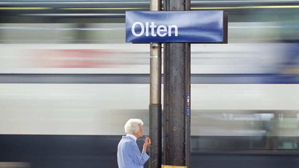 Bahnhofs-Schilt "Olten", im Hintergrund verschwommen ein Zug, der durchfährt, unten eine alte Frau, die auf dem Gleis steht. 