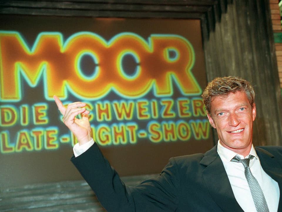 Ein Mann in Anzug und Krawatte sitzt mit erhobener Hand vor einer grossen Leinwand, auf der «Moor» steht.