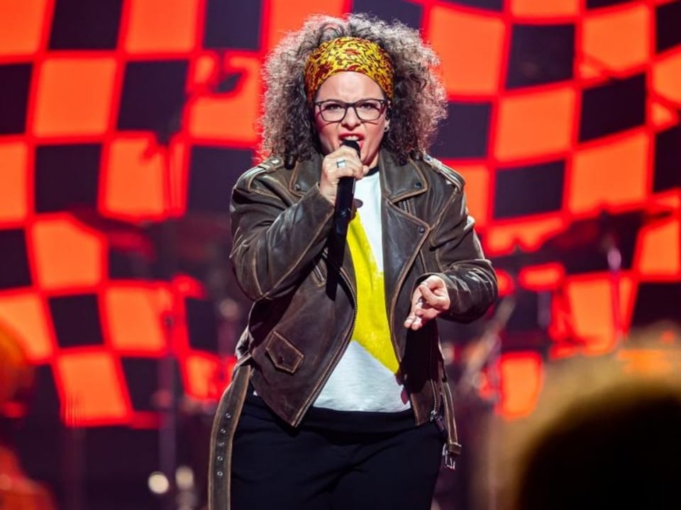 A woman with curly hair and glasses sings on stage.  She wears a brown leather jacket and a hair tie.