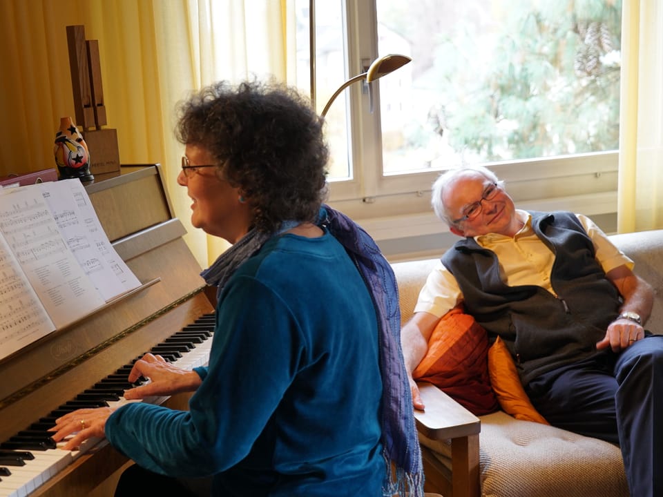 Frau spielt Klavier, Mann hört zu auf Sofa.