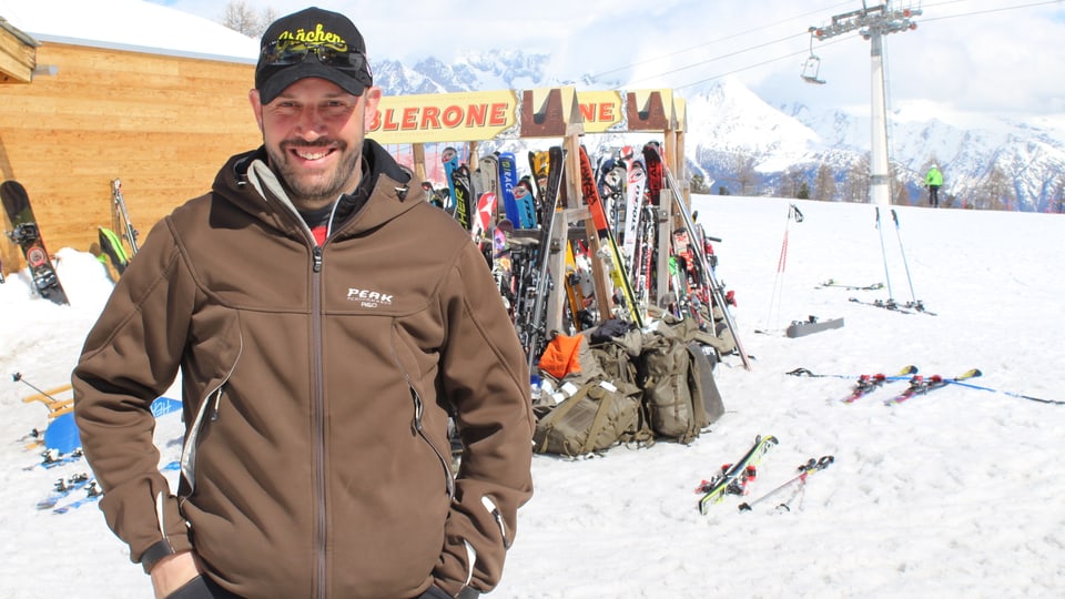 Ein Mann steht vor einem Skiständer und lacht in die Kamera. 