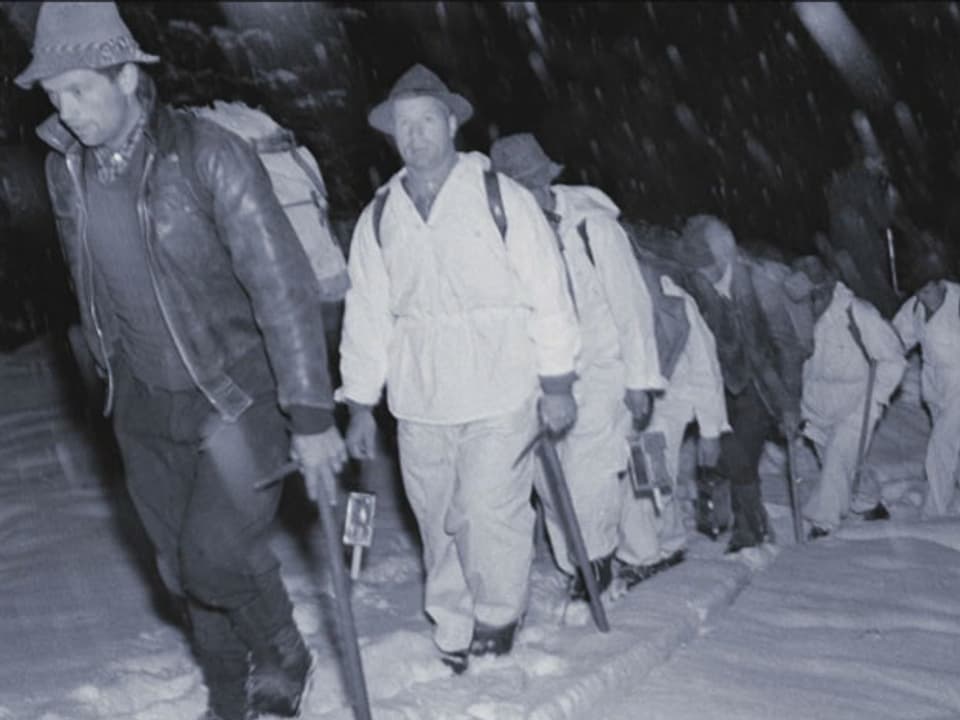 schwarzweiss: Eine Rettungskolonne mit Hüten und Eispickeln marschiert durch den Schnee. 