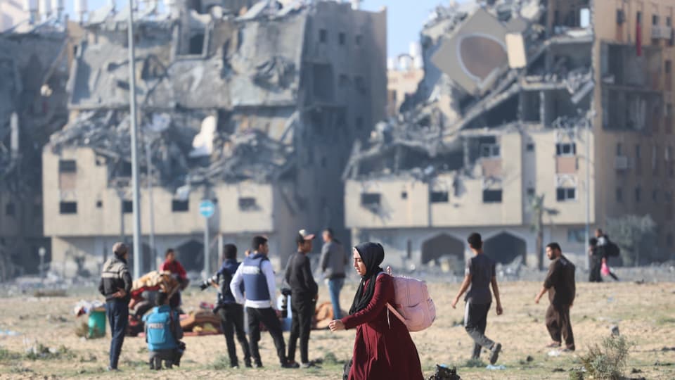 Menschenmenge steht einige Dutzend Meter von halb zerstörten Häusern entfernt, eine Frau läuft vorne im roten Kleid.