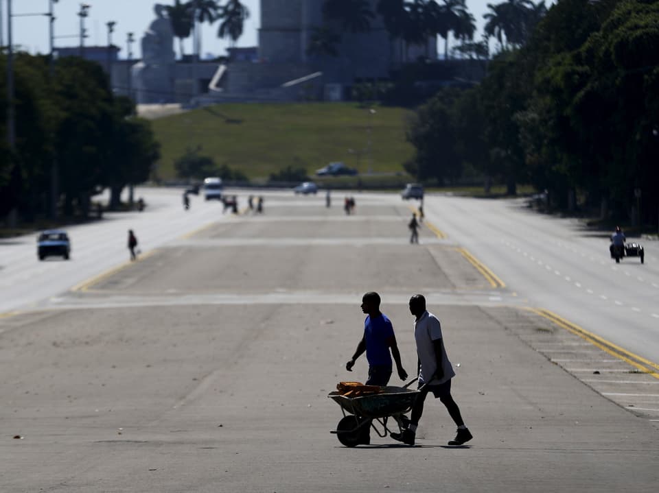 Zu sehen ist eine breite kubanische Strasse mit zwei Arbeitern, die eine Schubkarre stossen.