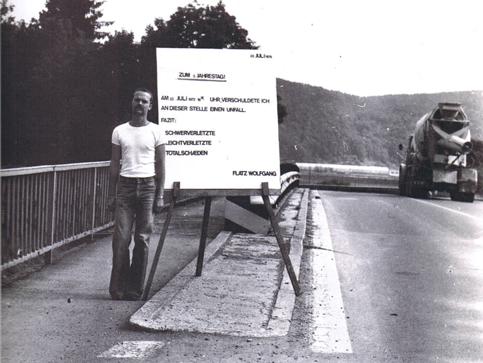 Schwarz-Weiss-Aufnahme: Flatz steht am Strassenrand neben einem Schild mit der Aufschrift «22. Juli 1975 zum 3. Jahrestag. Am 22. Juli 1972, 8.16 Uhr, verschuldete ich an dieser Stelle einen Unfall. Fazit: 3 Schwerverletzte, 2 Leichtverletzte, 2 Totalschäden. Flatz Wolfgang.»