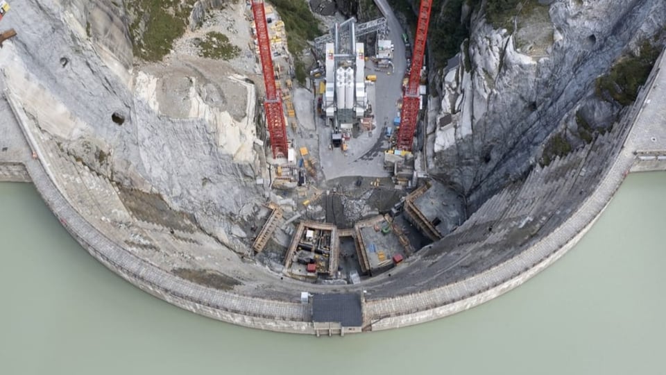 Eine Staumauer und eine dahinter liegende Baustelle sind von oben fotografiert worden.