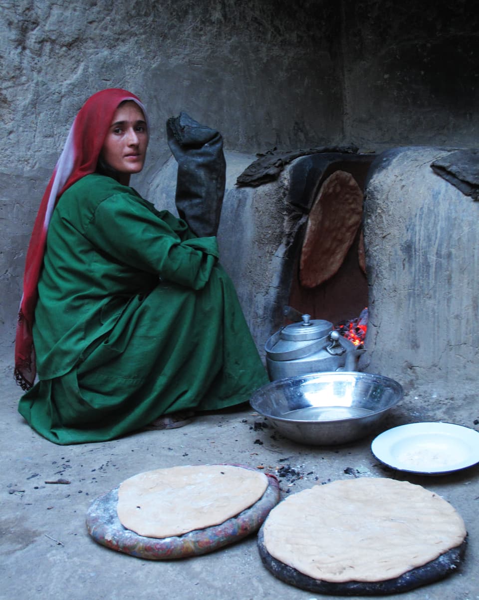 Ein Frau sitzt vor einem Steinofen. An der Innenwand klebt ein Fladenbrot, vor dem Ofen liegen weitere, ungebackene Fladenbrote auf Tellern bereit.