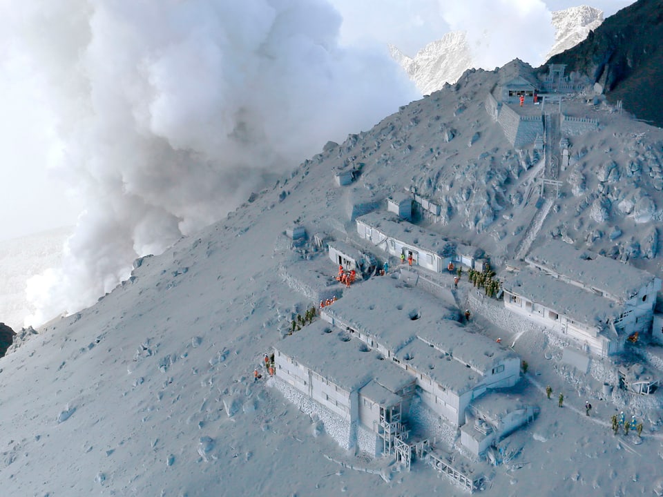 Die Unterkünfte und Hütten am Abhang des Berges sind wie die Landschaft mit Asche überzogen, im Hintergrund raucht der Vulkan Ontake.