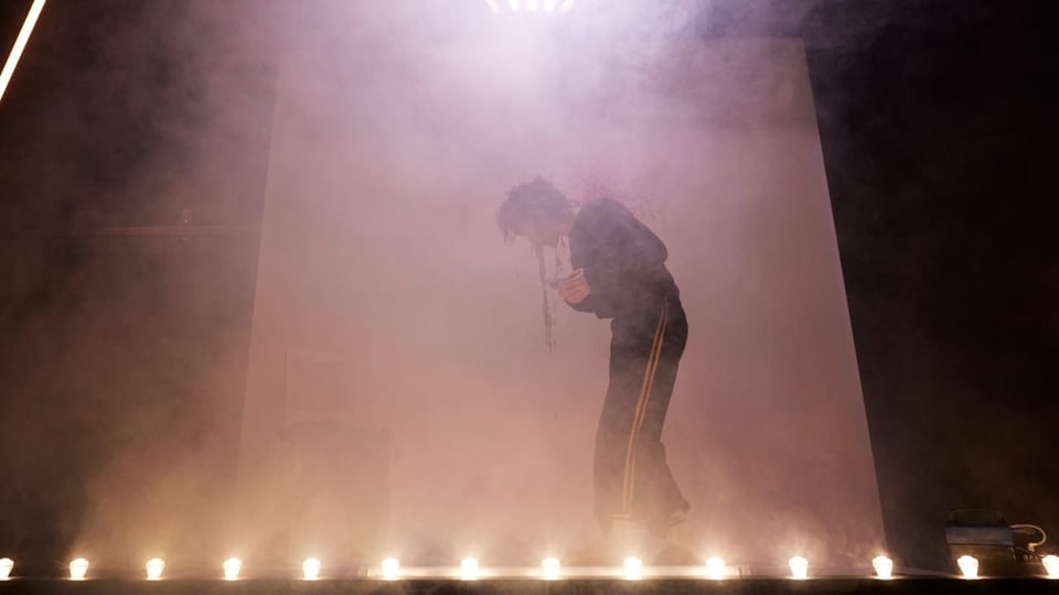 Eine Frau steht gebückt auf einer Bühne, sie verschwindet halb im Nebel.