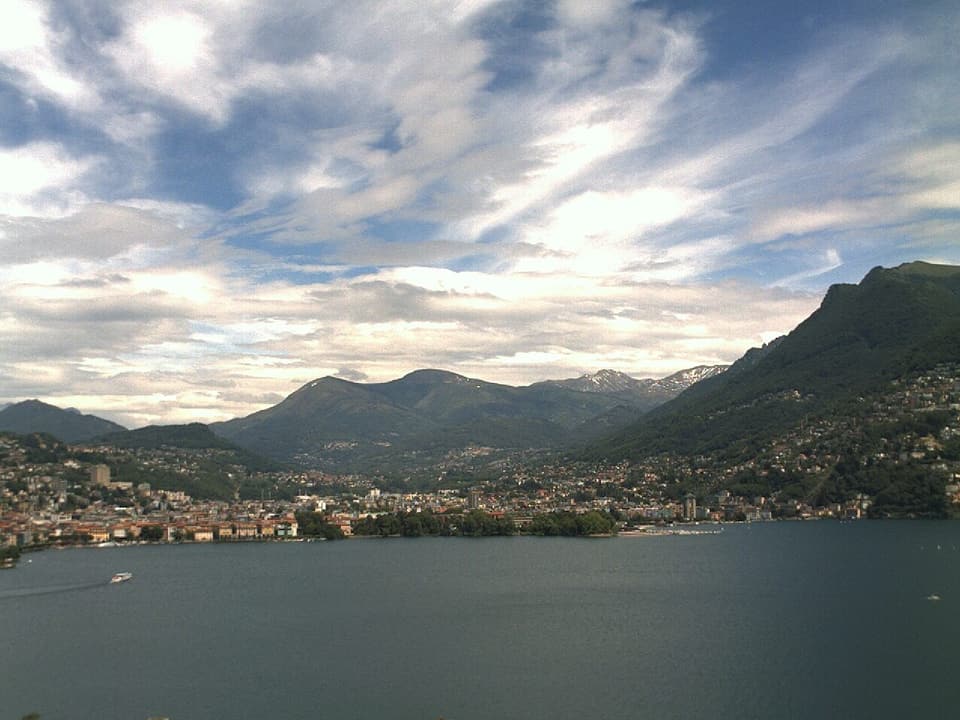 Im Vordergrund der Lago di Lugano mit einigen Schiffen darauf. Blick auf die Stadt Lugano und die nördlichen Berge. Der Himmel darüber weisst trotz Wolkenfelder viele blaue Flecken auf.
